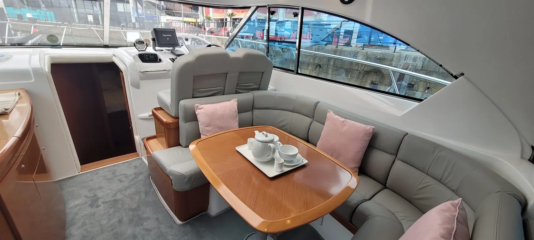 Luxury boat interior featuring a grey leather seating area with a wooden table, teapot, and cups, set against large windows.